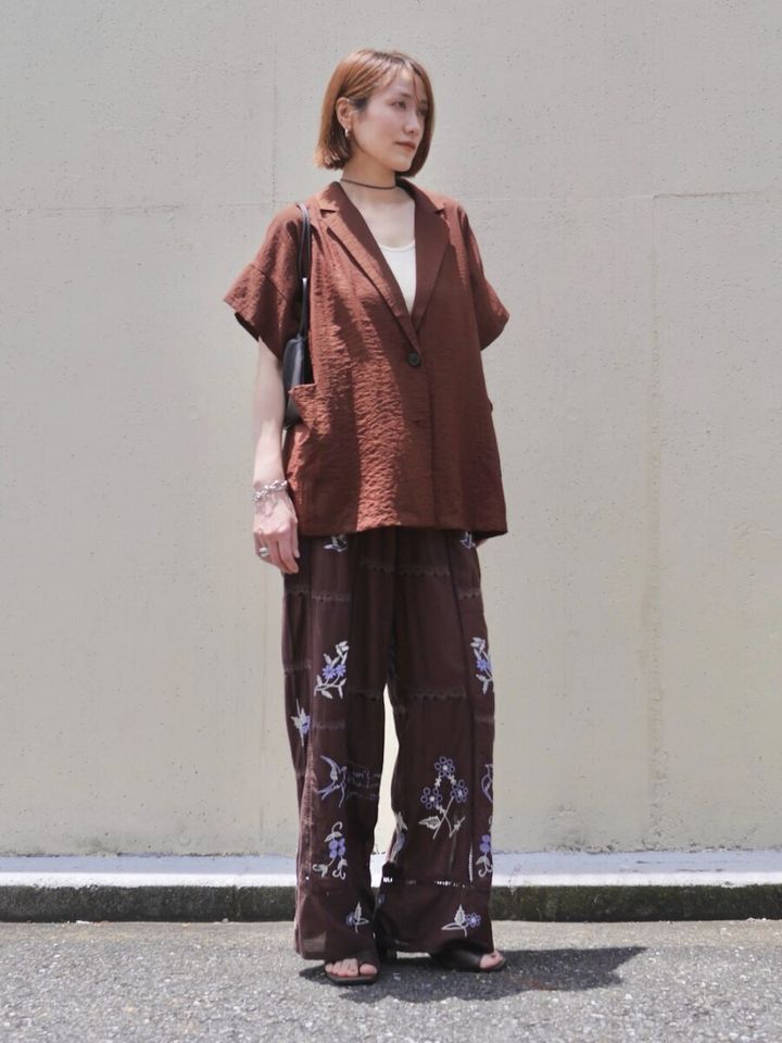 ブラウンの半袖ジャケットと刺繍入りトラウザーパンツを着用し、黒いミニバッグを持つ女性