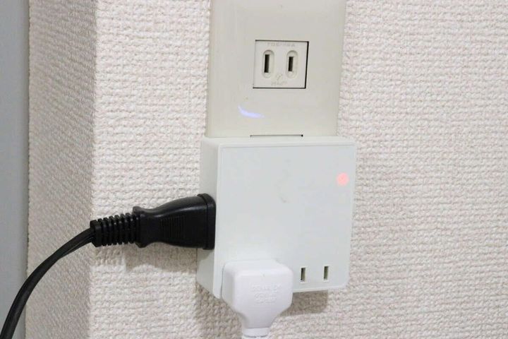 白い壁のコンセントに、白い4個口スクエアタップが差し込まれ、さらに黒い電源コードと白いプラグが接続されている