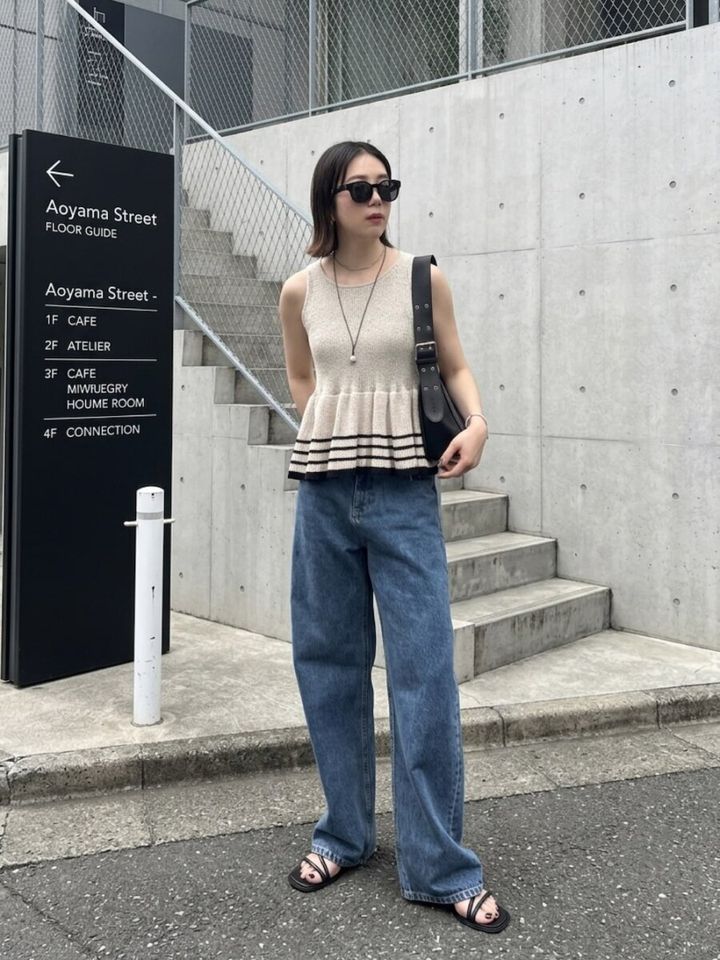 ベージュのペプラムニットとワイドデニム姿の女性が、屋外の階段前でポーズをとる