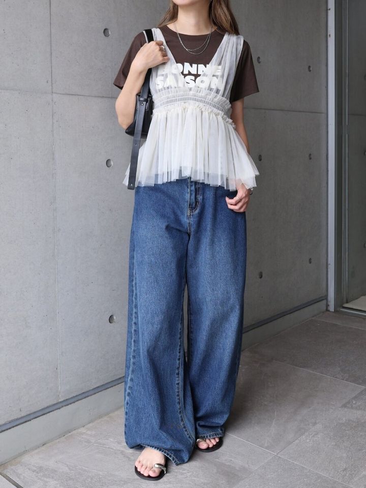 茶色Tシャツに白チュールビスチェ、ワイドデニム姿の女性が壁の前で立つ