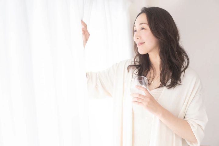 朝カーテンをあけて水を飲む女性