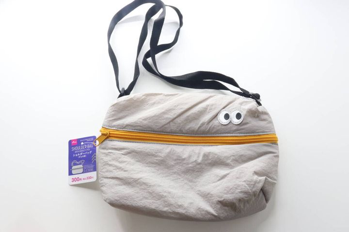 白い背景に置かれたグレーのギョロ目ショルダーバッグ。商品タグ付き。