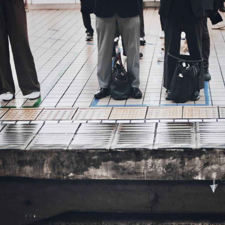 駅の向かい側のホームに並ぶ乗客たち。足元にはバッグが置かれ、ホームの端には点字ブロックが続いている。