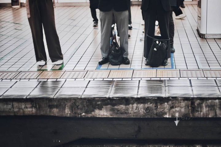 駅の向かい側のホームに並ぶ乗客たち。足元にはバッグが置かれ、ホームの端には点字ブロックが続いている。