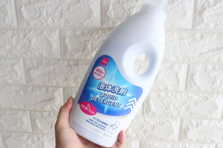 白い液体洗剤ボトルを右手が持ち上げている様子。白いレンガ調の壁を背景に