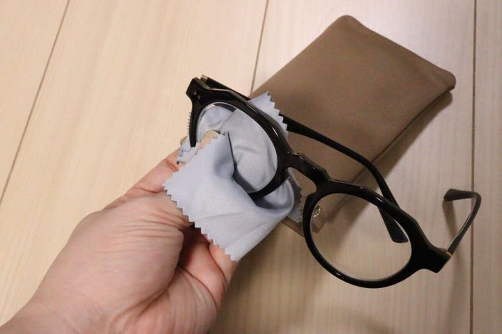 茶色のケースの横で黒縁メガネをグレーのメガネ拭きで拭く