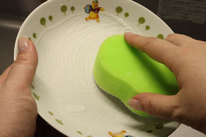 女性が緑色のキッチンスポンジで白いお皿を洗っている様子