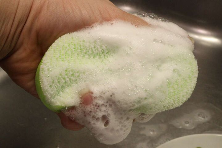 女性の手が洗剤で泡立てた緑色のキッチンスポンジを握っている