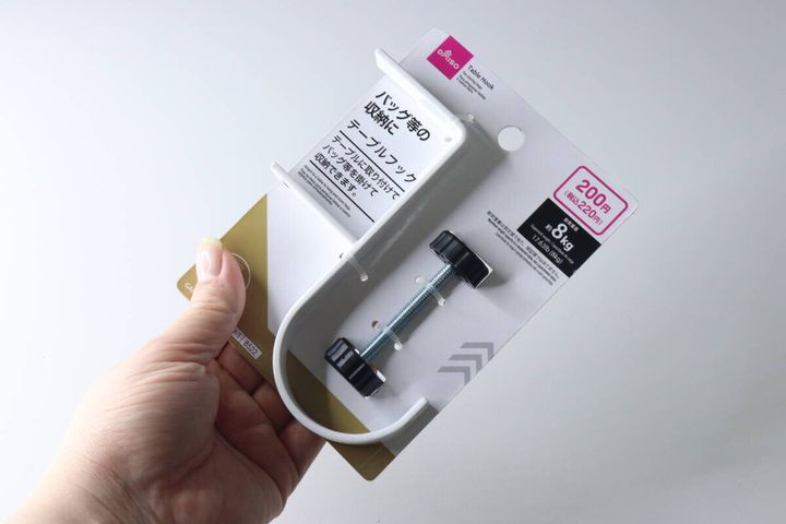 白い背景で、手がダイソーテーブルフックのパッケージを持っている