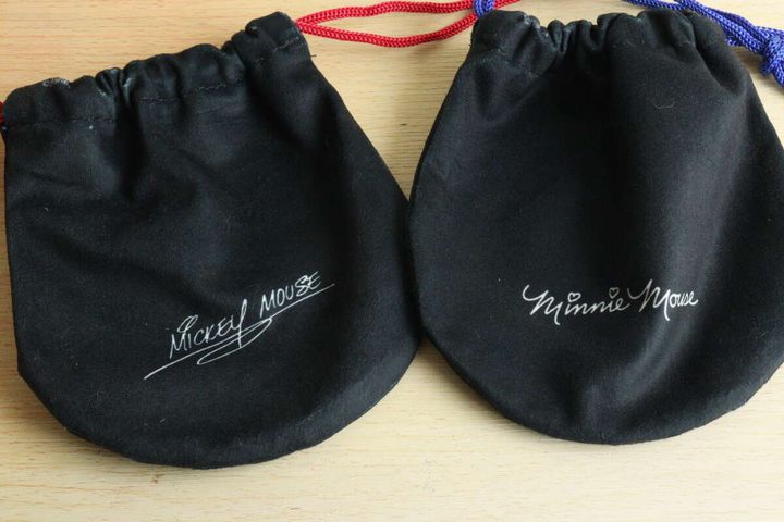 木目のテーブルに、ミッキーとミニーの巾着袋の裏面が並べられ、それぞれのキャラクターのサインが白い文字で書かれている。