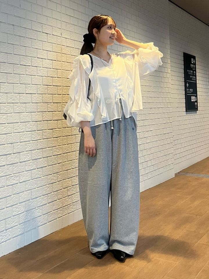 白いフリルブラウスとグレーのスウェットパンツを着用し、髪に手を当てる女性