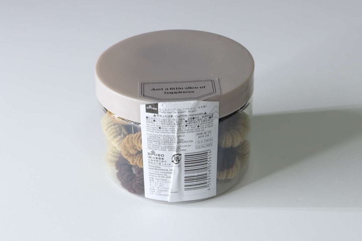 透明な円筒ケースに入ったブラウン系もこもこヘアゴム。商品ラベル付き。