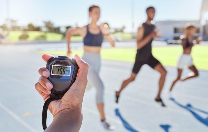 スポーツコーチがストップウォッチを使って選手のタイムを計測している。ゴールラインに向かって疾走し、記録を更新する選手たちの姿がぼやけて見える。スポーツイベントでマラソンのタイムを計測するストップウォッチ。