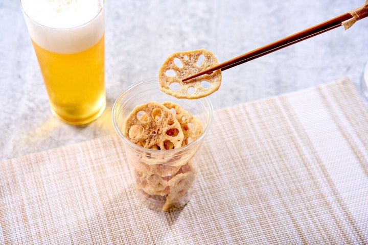 透明なカップに入ったレンコンチップを箸でつまんでいる様子。奥にはビールグラスが置かれている。