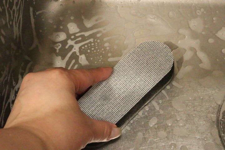 女性の手がダイソーのスポンジの白い面で泡立ったシンクを掃除している