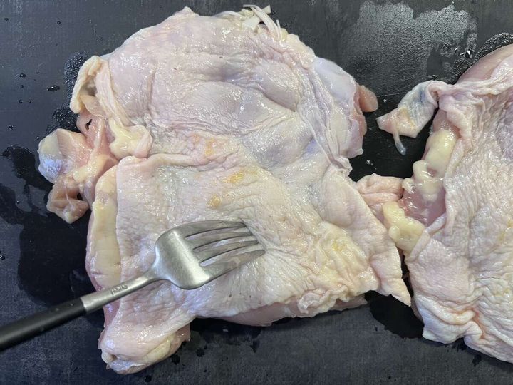 黒いまな板の上で鶏もも肉の皮目にフォークで穴を開けている手元