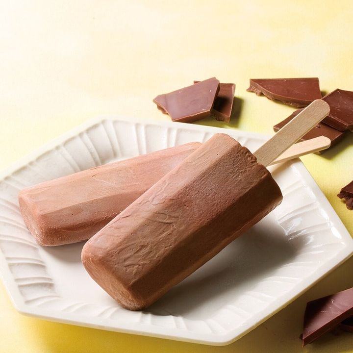 黄色い背景の白い皿に置かれたチョコアイスバー2本と砕けた板チョコ