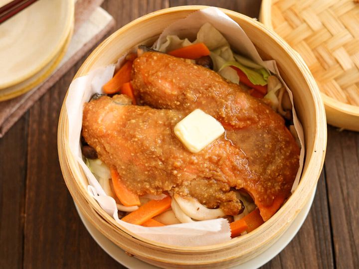 蒸し上がったせいろの中の鮭と野菜の上にバターが乗せられ、奥にはせいろの蓋が置かれている。木製のテーブルの上で、全体が明るく写っている