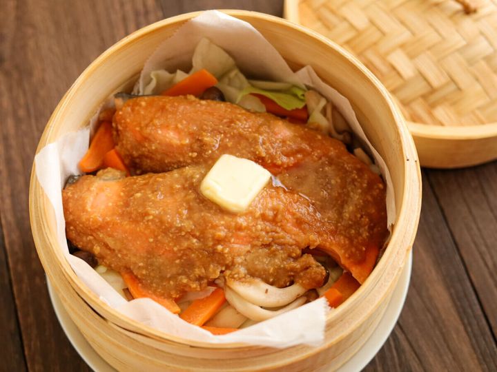 蒸し上がったせいろの中の鮭と野菜の上にバターが乗せられ、奥にはせいろの蓋が置かれている。木製のテーブルの上
