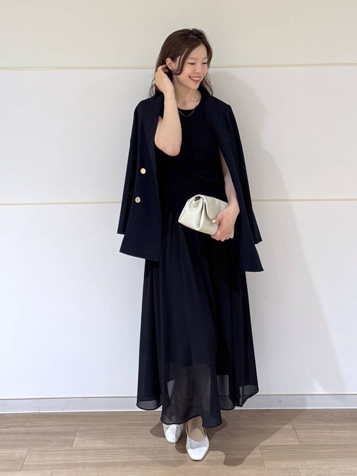 黒いノースリーブワンピにジャケットを羽織り白いクラッチバッグを持つ女性