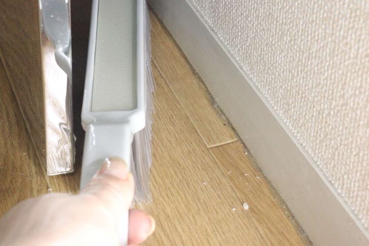 女性の手が、家具と壁の狭い隙間にほうきを差し込んで掃除している様子