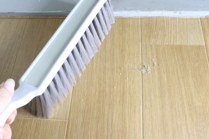 女性の手が、フローリングの床に散らばったゴミをほうきで掃いている様子