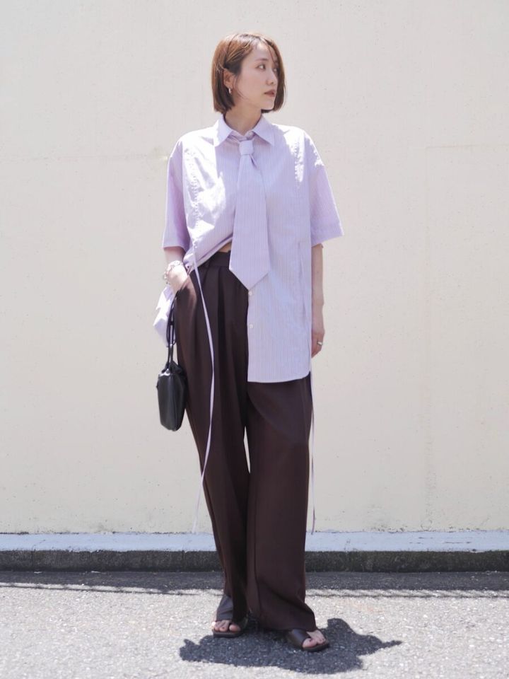 淡いパープルのネクタイ付きシャツとブラウンのワイドパンツを着用し、黒いミニバッグを持った女性が屋外で立っている