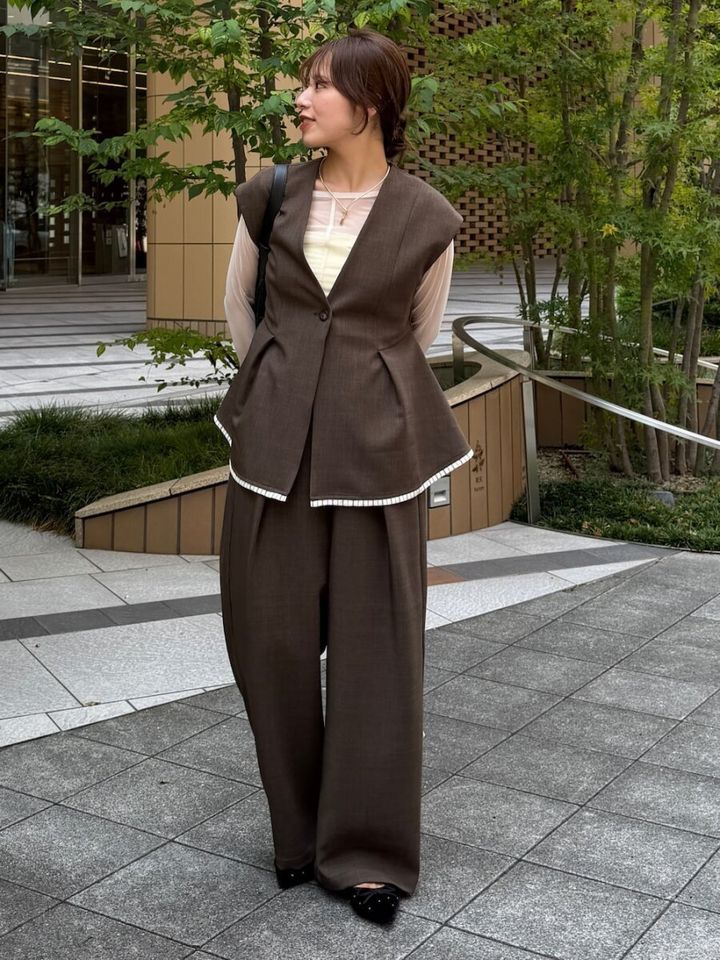ブラウンのペプラムベストとワイドパンツのセットアップを着用し、屋外で横を向いて立っている女性