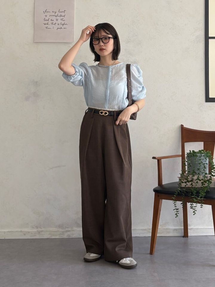 淡いブルーのパフスリーブブラウスとブラウンのワイドパンツを着用し、眼鏡をかけた女性が髪に手を当てている
