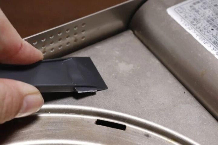 ガスコンロの隙間に使い捨ての黒いヘラを差し込み汚れをかき出す手元