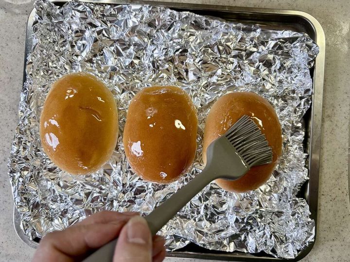 アルミホイルを敷いた天板に並んだロールパン3個に、刷毛でサラダ油を塗っている手元