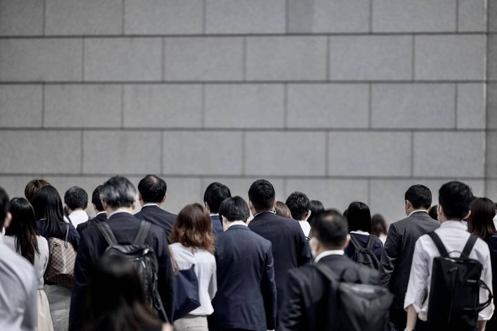 スーツ姿の人々が大勢集まり、建物の前に並んでいる様子。