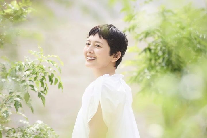 明るい日差しの中、白いブラウスを着たショートヘアの女性が、柔らかな笑顔で上を見上げているポートレート。背景の緑が爽やかな印象を与えています。