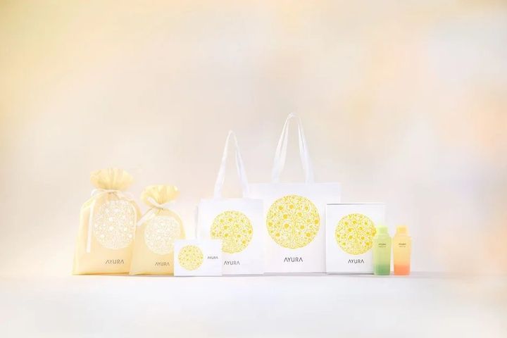 AYURAブランドの化粧品やギフトセットのパッケージ、トートバッグ、巾着袋、そして小瓶が並べられた画像です。白と黄色を基調とした統一感のあるデザインが特徴で、清潔感と上品さを感じさせます。
