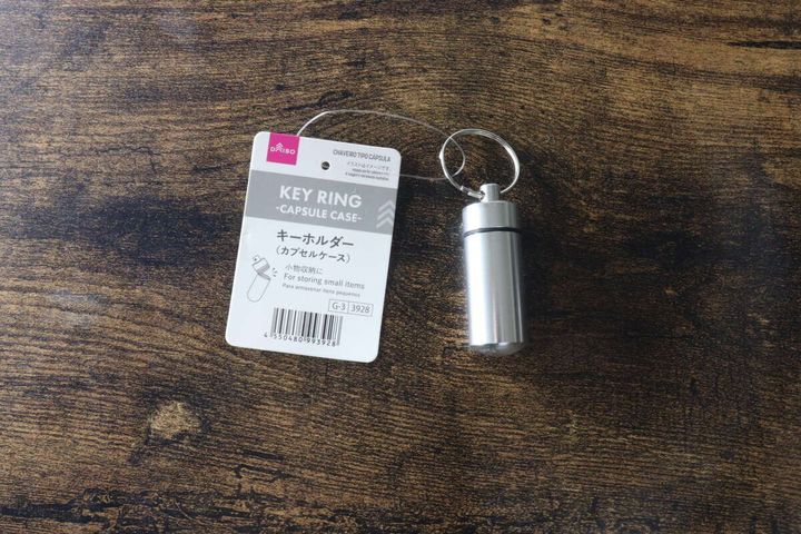 木目調のテーブルに置かれた、パッケージ付きの銀色カプセルケースキーホルダー