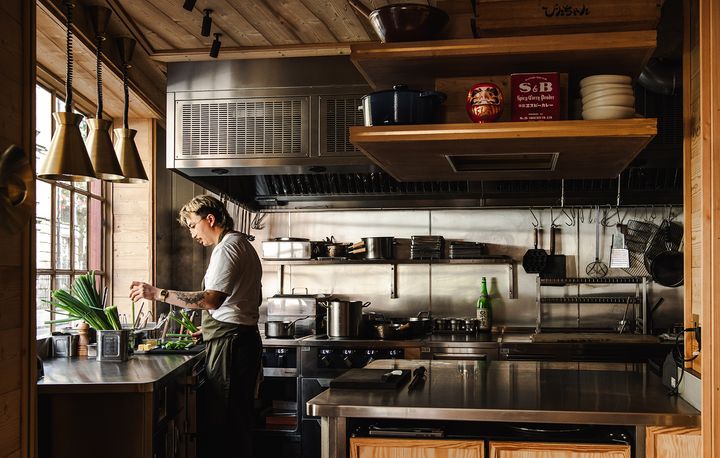 木の棚と銅色のペンダントライトがあるオープンキッチンで、料理の仕込みをするロビン・コスゲ