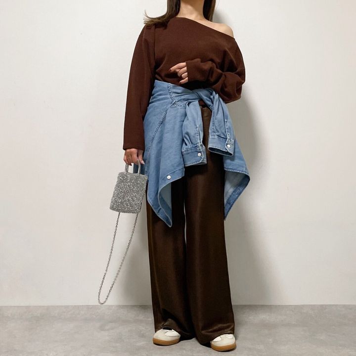 焦茶色のオフショルダーニットとパンツを着用し、腰にデニムシャツを巻いた女性