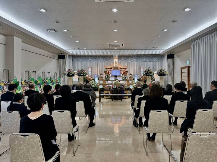 葬儀会場で、祭壇に向かって椅子に座る参列者たちの後ろ姿。