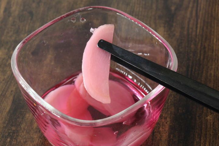 透明なガラスの器に入ったピンク色の新生姜スライスを黒いトングでつまみ上げている