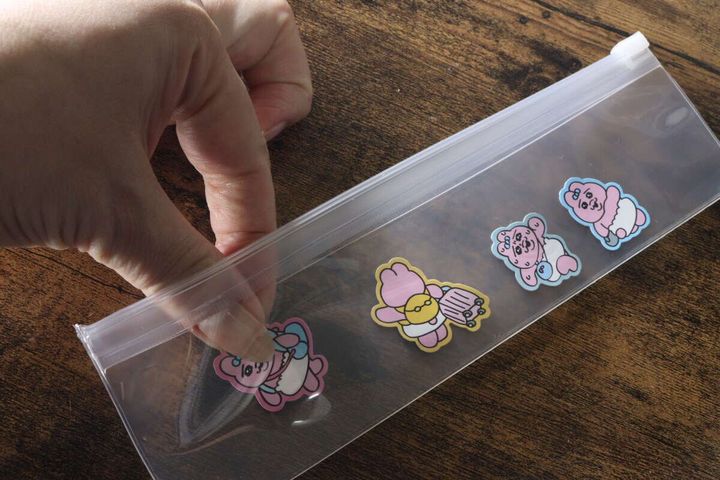 女性の手が、細長いクリアポーチにクマのイラストが描かれた缶バッジを入れている様子