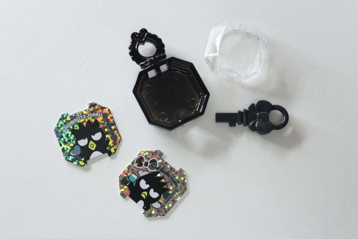 分解されたジュエルキーホルダー本体と透明なフタ、鍵、2種類のバッドばつ丸シートが並べられている