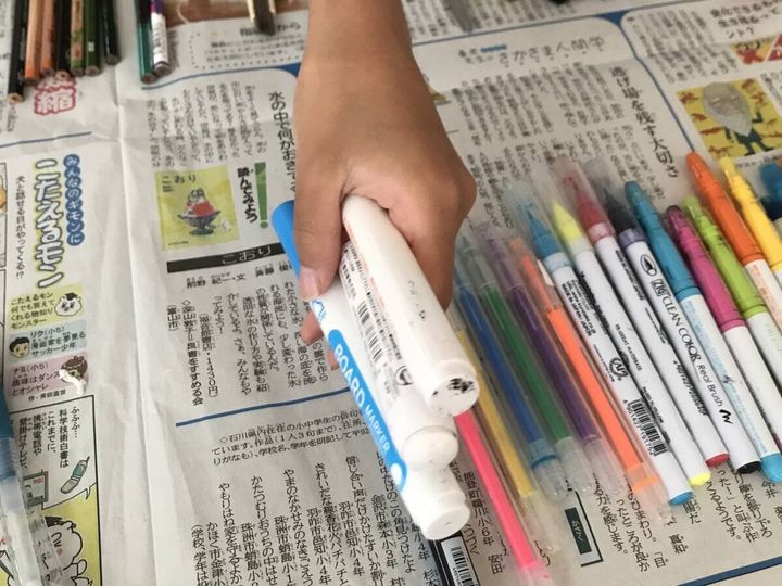 新聞紙の上に広げられた色とりどりのペンと白いマーカーを掴む子どもの手