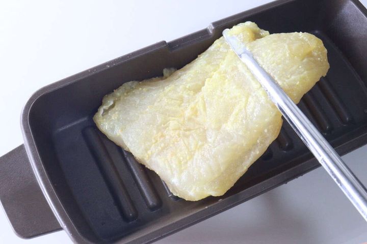 茶色い調理器具に乗った魚の切り身をステンレス製の「箸トング」で掴んでいる