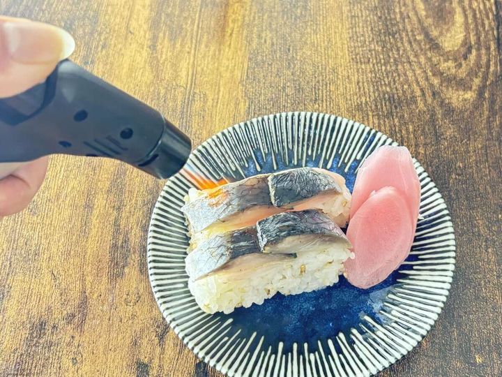 手がダイソーのガスバーナーを使いお皿に乗った鯖寿司を炙っている