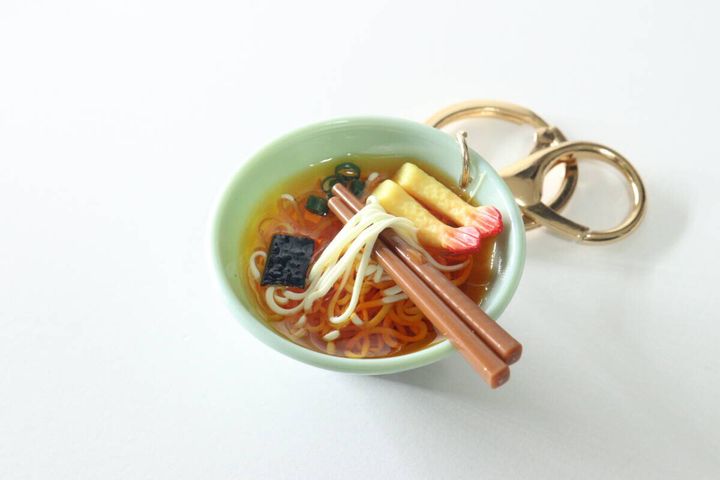 白い背景に置かれた、箸で麺を持ち上げたデザインのうどんキーホルダー。エビ天やネギもリアルに再現。