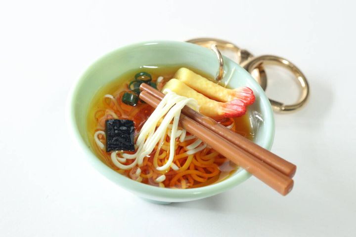 白い背景に置かれたうどんキーホルダーのアップ。麺や具材、透き通ったスープのリアルな質感がわかる。