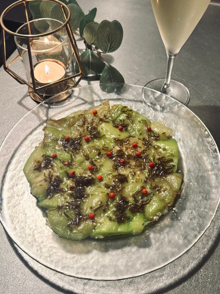 ガラス皿に茄子を潰し広げ、塩昆布を刻んだソース、ピンクペッパーをのせたおつまみと、背景にシャンパングラス、キャンドル、グリーン