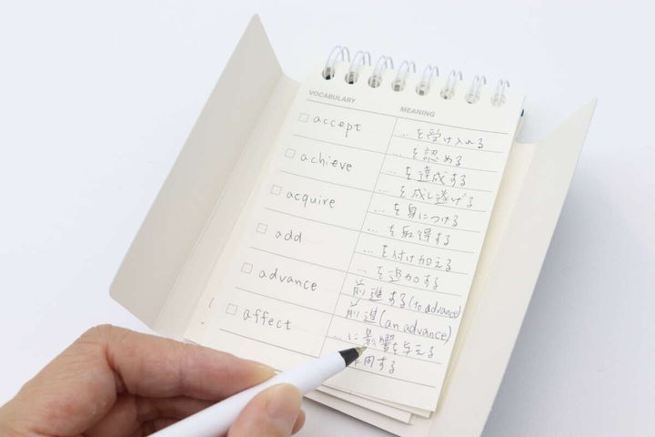 白いペンを持った手が、単語帳のページに英単語と意味を書き込んでいる様子