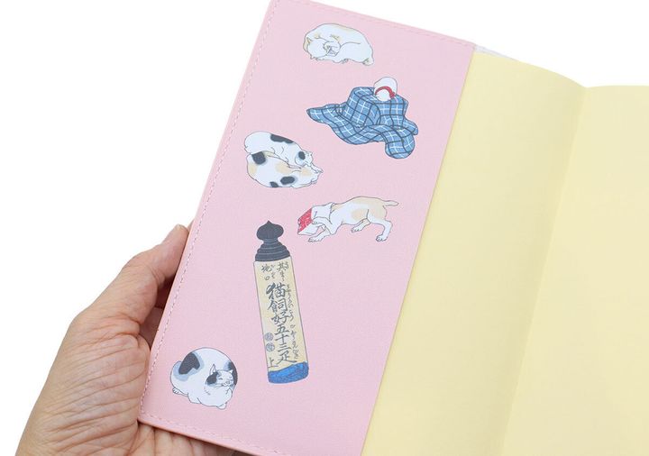 ピンクのレザー調カバーを開き、内側の折り返し部分に描かれた猫のイラストと題字が見える。