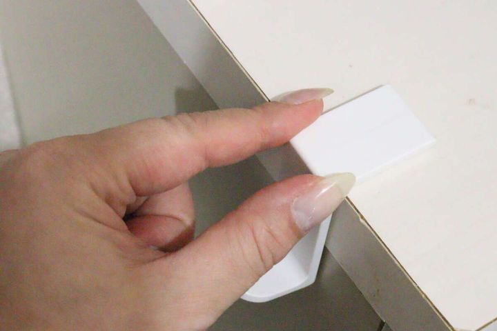 白いL字型コーナーフックを白い家具の角に当てて貼り付けようとしている手元